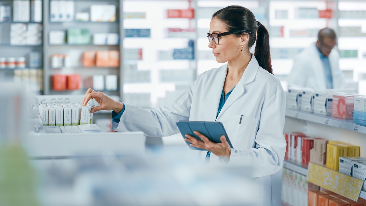 Pharmacy Drugstore: Beautiful Caucasian Pharmacist Uses Digital Tablet Computer, Checks Inventory of Medicine, Drugs, Vitamins, Health Care Products on a Shelf. Professional Pharmacist in Pharma Store